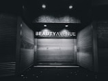 An urban alley scene features a closed storefront with metal shutters and a sign above reading 'Beauty Avenue.' The area is dimly lit with overhead lights creating a stark contrast against the dark surroundings.