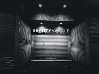 An urban alley scene features a closed storefront with metal shutters and a sign above reading 'Beauty Avenue.' The area is dimly lit with overhead lights creating a stark contrast against the dark surroundings.