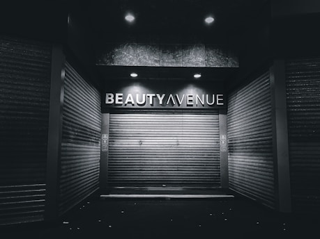 An urban alley scene features a closed storefront with metal shutters and a sign above reading 'Beauty Avenue.' The area is dimly lit with overhead lights creating a stark contrast against the dark surroundings.