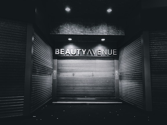 An urban alley scene features a closed storefront with metal shutters and a sign above reading 'Beauty Avenue.' The area is dimly lit with overhead lights creating a stark contrast against the dark surroundings.