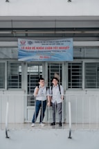 Two young men with backpacks stand in front of a building. The building has a banner written in Vietnamese that appears to be related to graduation or an academic event. The door and windows behind them are barred. They are standing on a small balcony with railings.