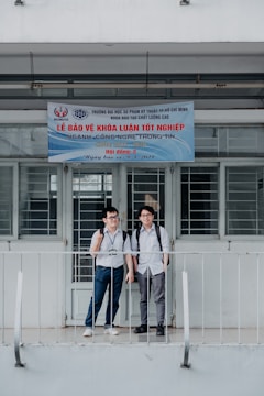 Two young men with backpacks stand in front of a building. The building has a banner written in Vietnamese that appears to be related to graduation or an academic event. The door and windows behind them are barred. They are standing on a small balcony with railings.