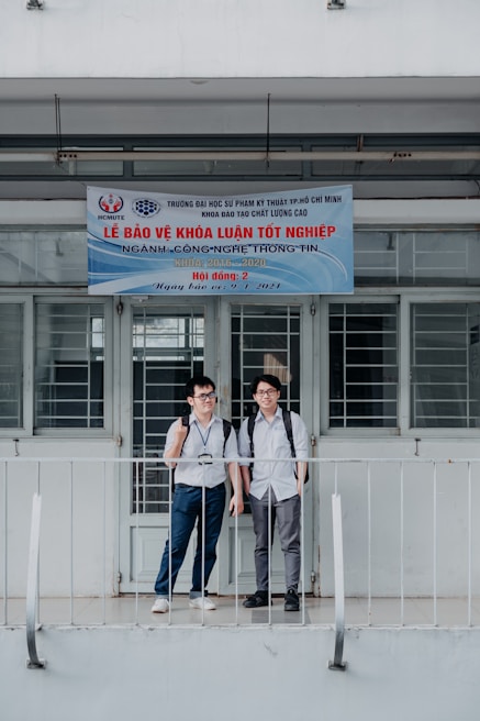 Two young men with backpacks stand in front of a building. The building has a banner written in Vietnamese that appears to be related to graduation or an academic event. The door and windows behind them are barred. They are standing on a small balcony with railings.