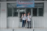 Two young men stand in front of a building entrance with signs overhead, one making a peace sign with his fingers. Both are wearing casual shirts and backpacks, suggesting a school or academic setting. A metal railing is in front of them, and the building appears to have a modest, institutional design with barred windows.