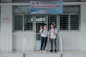 Two young men stand in front of a building entrance with signs overhead, one making a peace sign with his fingers. Both are wearing casual shirts and backpacks, suggesting a school or academic setting. A metal railing is in front of them, and the building appears to have a modest, institutional design with barred windows.