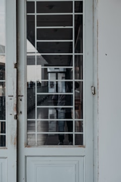 A close-up view of a partially open door with a large glass panel featuring a white grid pattern. There is a reflection of a person standing behind the door, creating an intriguing visual depth. The hallway seen through the glass appears modern and well-lit, with additional doors and windows visible in the background.