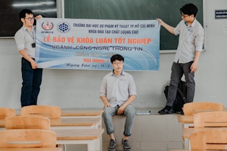 Three individuals are in a classroom setting, with two standing and holding a banner while another sits on a ledge. The banner is written in Vietnamese with text related to a thesis defense event at a university. The room contains several wooden chairs and a blackboard can be seen behind them.
