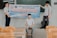 Three individuals are in a classroom setting, with two standing and holding a banner while another sits on a ledge. The banner is written in Vietnamese with text related to a thesis defense event at a university. The room contains several wooden chairs and a blackboard can be seen behind them.