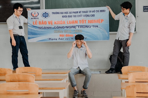Three individuals are in a classroom setting, with two standing on desks holding a large banner and one seated below them adjusting glasses. The room has wooden desks and chairs, and a green chalkboard is visible in the background.