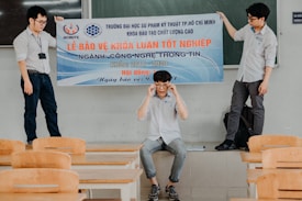 Three individuals are in a classroom setting, with two standing on desks holding a large banner and one seated below them adjusting glasses. The room has wooden desks and chairs, and a green chalkboard is visible in the background.