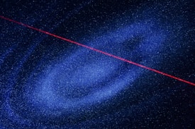 A spiral galaxy with numerous stars scattered throughout. A vivid red laser beam cuts diagonally across the image, contrasting against the deep blue and black starry background.
