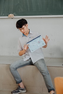 A person with short dark hair and glasses is sitting on a step in a classroom setting, holding a transparent folder containing papers. They are wearing a light-colored shirt and jeans, and there is a chalkboard in the background.