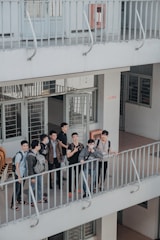 A group of young individuals stand together on a balcony, engaged in conversation and displaying various expressions. The environment appears to be a multi-level building with open corridors and railings. They are casually dressed, and some carry backpacks, suggesting a school or college setting.