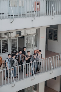 A group of young individuals stand together on a balcony, engaged in conversation and displaying various expressions. The environment appears to be a multi-level building with open corridors and railings. They are casually dressed, and some carry backpacks, suggesting a school or college setting.