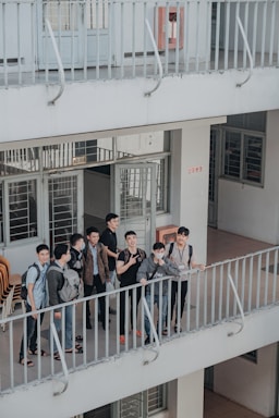 A group of young individuals stand together on a balcony, engaged in conversation and displaying various expressions. The environment appears to be a multi-level building with open corridors and railings. They are casually dressed, and some carry backpacks, suggesting a school or college setting.