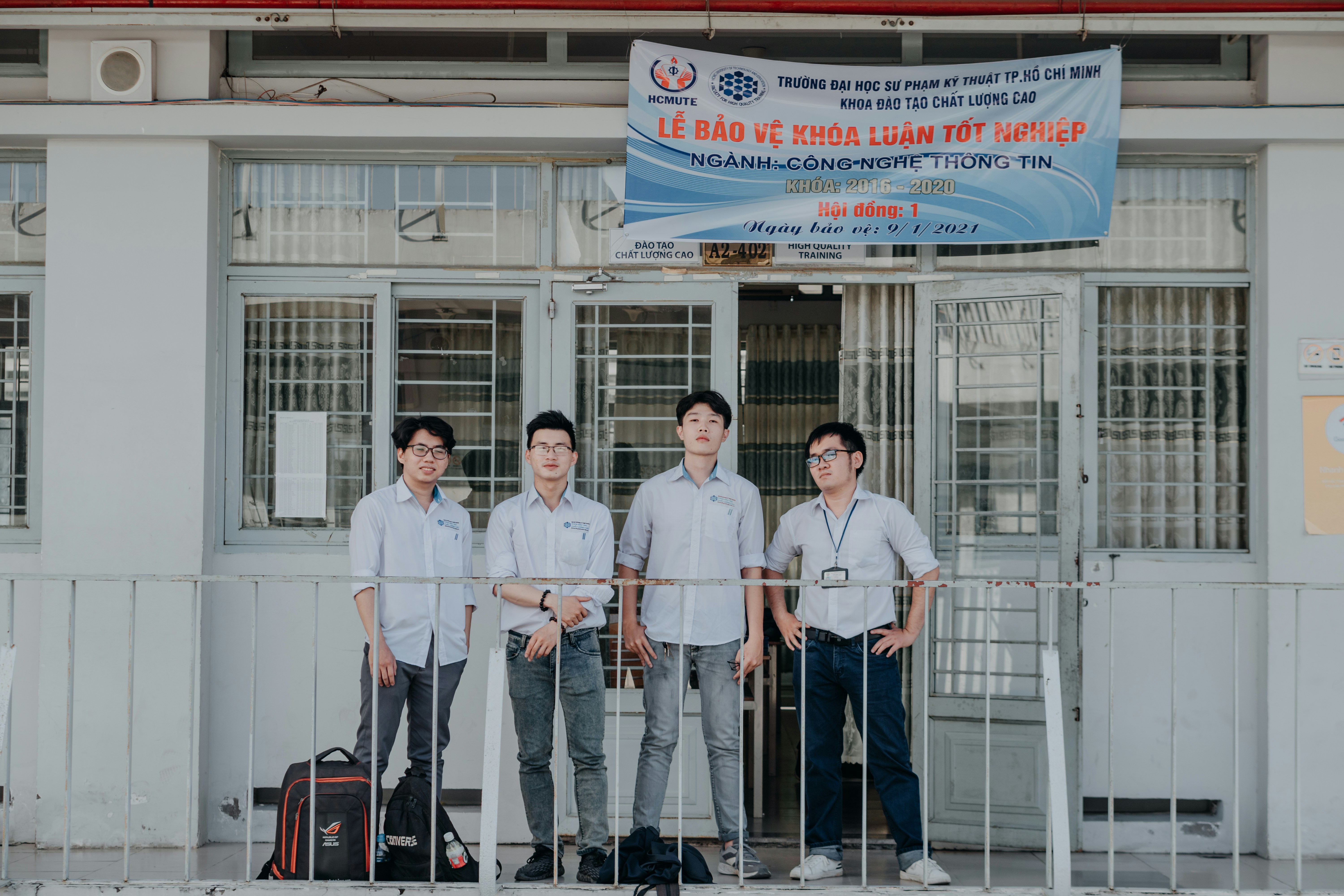 Four individuals stand in front of a building wearing formal button-down shirts with name tags. They are positioned near a sign above the door with details in Vietnamese. The setting appears academic or professional, likely related to a university or educational institution.