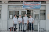 Four individuals stand in front of a building wearing formal button-down shirts with name tags. They are positioned near a sign above the door with details in Vietnamese. The setting appears academic or professional, likely related to a university or educational institution.