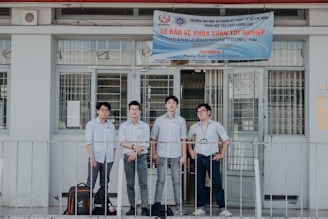 Four individuals stand in front of a building wearing formal button-down shirts with name tags. They are positioned near a sign above the door with details in Vietnamese. The setting appears academic or professional, likely related to a university or educational institution.