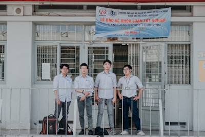 Four individuals stand in front of a building wearing formal button-down shirts with name tags. They are positioned near a sign above the door with details in Vietnamese. The setting appears academic or professional, likely related to a university or educational institution.