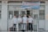 Four individuals stand in front of a building wearing formal button-down shirts with name tags. They are positioned near a sign above the door with details in Vietnamese. The setting appears academic or professional, likely related to a university or educational institution.