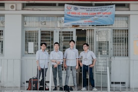 Four individuals stand in front of a building wearing formal button-down shirts with name tags. They are positioned near a sign above the door with details in Vietnamese. The setting appears academic or professional, likely related to a university or educational institution.