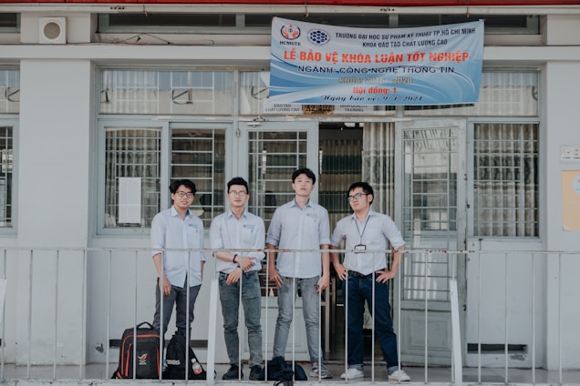 Four individuals stand in front of a building wearing formal button-down shirts with name tags. They are positioned near a sign above the door with details in Vietnamese. The setting appears academic or professional, likely related to a university or educational institution.