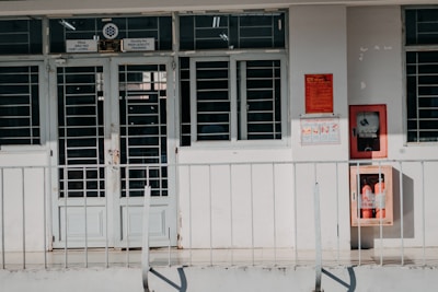 A building entrance with metal barred doors and windows, featuring signs indicating it belongs to a faculty for high-quality training. The facade is white, and there are red fire extinguishers and safety notices mounted on the wall next to the entrance.