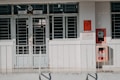 A building entrance with metal barred doors and windows, featuring signs indicating it belongs to a faculty for high-quality training. The facade is white, and there are red fire extinguishers and safety notices mounted on the wall next to the entrance.