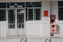 A building entrance with metal barred doors and windows, featuring signs indicating it belongs to a faculty for high-quality training. The facade is white, and there are red fire extinguishers and safety notices mounted on the wall next to the entrance.
