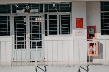 A building entrance with metal barred doors and windows, featuring signs indicating it belongs to a faculty for high-quality training. The facade is white, and there are red fire extinguishers and safety notices mounted on the wall next to the entrance.
