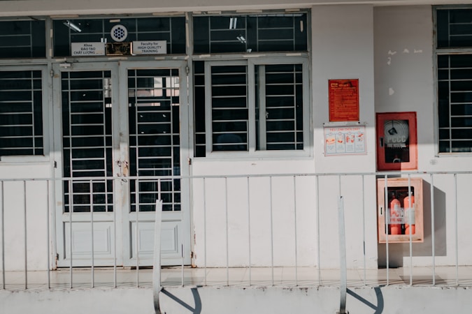 A building entrance with metal barred doors and windows, featuring signs indicating it belongs to a faculty for high-quality training. The facade is white, and there are red fire extinguishers and safety notices mounted on the wall next to the entrance.