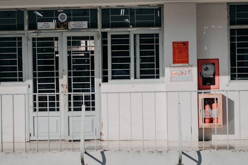 A building entrance with metal barred doors and windows, featuring signs indicating it belongs to a faculty for high-quality training. The facade is white, and there are red fire extinguishers and safety notices mounted on the wall next to the entrance.