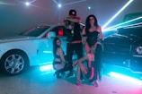 In a dimly lit setting accentuated by vibrant neon light beams, a fashionable group poses confidently between two luxury cars, a white Rolls Royce and a black Mercedes-Benz. They are dressed in stylish, revealing clubwear, exuding an aura of opulence and flair. The shiny surfaces of the cars reflect the ambient lighting, creating a dynamic, high-energy atmosphere.