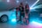 In a dimly lit setting accentuated by vibrant neon light beams, a fashionable group poses confidently between two luxury cars, a white Rolls Royce and a black Mercedes-Benz. They are dressed in stylish, revealing clubwear, exuding an aura of opulence and flair. The shiny surfaces of the cars reflect the ambient lighting, creating a dynamic, high-energy atmosphere.