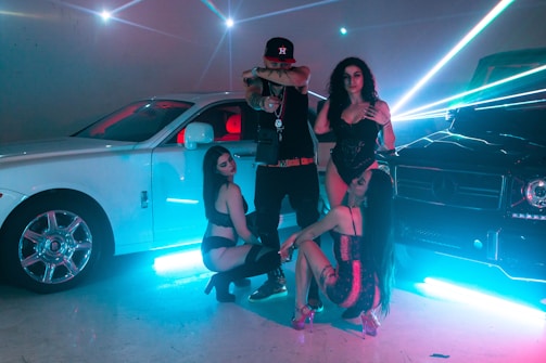 In a dimly lit setting accentuated by vibrant neon light beams, a fashionable group poses confidently between two luxury cars, a white Rolls Royce and a black Mercedes-Benz. They are dressed in stylish, revealing clubwear, exuding an aura of opulence and flair. The shiny surfaces of the cars reflect the ambient lighting, creating a dynamic, high-energy atmosphere.