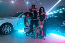 In a dimly lit setting accentuated by vibrant neon light beams, a fashionable group poses confidently between two luxury cars, a white Rolls Royce and a black Mercedes-Benz. They are dressed in stylish, revealing clubwear, exuding an aura of opulence and flair. The shiny surfaces of the cars reflect the ambient lighting, creating a dynamic, high-energy atmosphere.