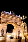 An elaborate architectural structure featuring an archway adorned with sculptures and intricate carvings. The entrance is illuminated with bright lights, and figures of horses and a chariot are positioned on top. The word 'Europe' is displayed in bold, bright lettering. Many people are gathered around the entrance under the night sky.