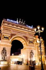 An elaborate architectural structure featuring an archway adorned with sculptures and intricate carvings. The entrance is illuminated with bright lights, and figures of horses and a chariot are positioned on top. The word 'Europe' is displayed in bold, bright lettering. Many people are gathered around the entrance under the night sky.