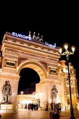 An elaborate architectural structure featuring an archway adorned with sculptures and intricate carvings. The entrance is illuminated with bright lights, and figures of horses and a chariot are positioned on top. The word 'Europe' is displayed in bold, bright lettering. Many people are gathered around the entrance under the night sky.