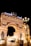 An elaborate architectural structure featuring an archway adorned with sculptures and intricate carvings. The entrance is illuminated with bright lights, and figures of horses and a chariot are positioned on top. The word 'Europe' is displayed in bold, bright lettering. Many people are gathered around the entrance under the night sky.