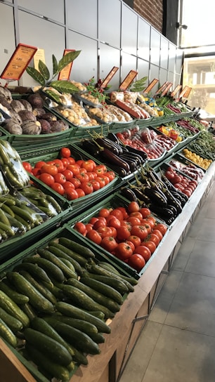 A well-organized display of fresh vegetables in a market setting, including baskets of cucumbers, tomatoes, eggplants, and various root vegetables. Bright labels and signs are visible above each section, and natural light illuminates the produce, enhancing their vibrant colors.