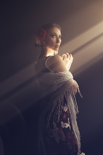 A woman stands in a dramatic light, wearing a floral dress and a fringed shawl. Her hair is adorned with a large red flower, and the lighting creates a soft, ethereal glow around her. She is posed elegantly, with hands slightly raised and turned towards the light.