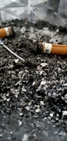 Cigarette butts and ash are scattered across a surface, creating a gritty, textured appearance. The image features partially smoked cigarettes with visible remnants of tobacco.