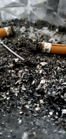 Cigarette butts and ash are scattered across a surface, creating a gritty, textured appearance. The image features partially smoked cigarettes with visible remnants of tobacco.