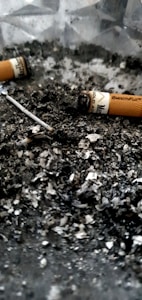 Cigarette butts and ash are scattered across a surface, creating a gritty, textured appearance. The image features partially smoked cigarettes with visible remnants of tobacco.