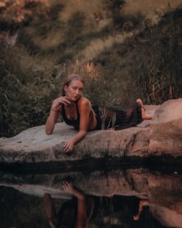 A person is lounging on a rock near a calm body of water, surrounded by lush greenery. Their reflection is visible in the water, creating a serene and introspective scene.