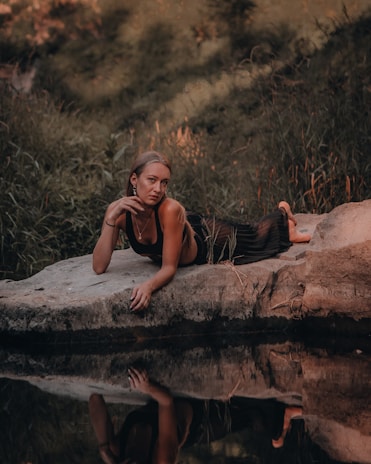 A person is lounging on a rock near a calm body of water, surrounded by lush greenery. Their reflection is visible in the water, creating a serene and introspective scene.