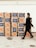 A person wearing a mask walks past a stack of cardboard boxes labeled 'Love Box' and adorned with Home Depot logos. The boxes are neatly organized against a light-colored wall.