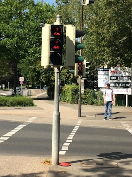 Several traffic lights are mounted on a pole at a pedestrian crossing with red signals indicating stop. A person is standing on the sidewalk near the crossing. Lush green trees are in the background along with signage near the sidewalk.