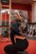 A woman is kneeling on a padded surface in a gym setting, wearing headphones and a decorative face mask. She has long, wavy hair tied back and is dressed in athletic wear, including a cropped hoodie with the word 'fitness' visible. The background features gym equipment and a red carpeted floor.
